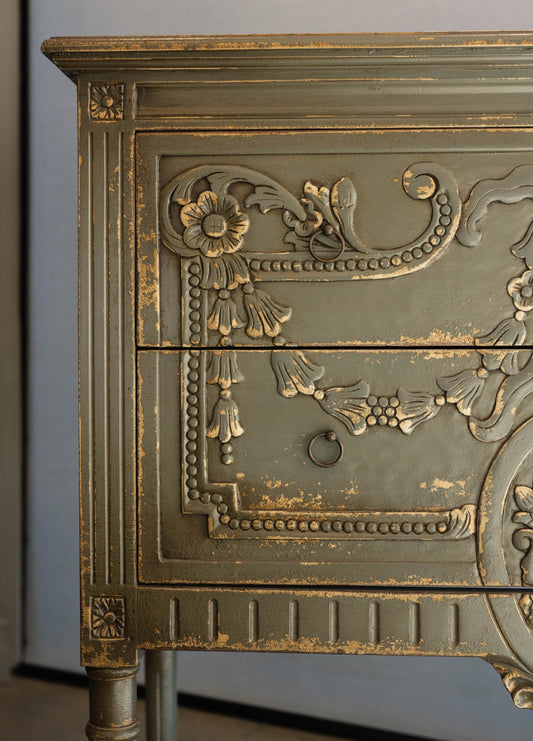 The unique and stunning one of a kind Daphne Sideboard boasts intricate carvings and distressed pale olive/grey painted finish. Crafted from mahogany and featuring two drawers, this versatile piece is perfect for a living or dining room in even in a bedroom. Detail