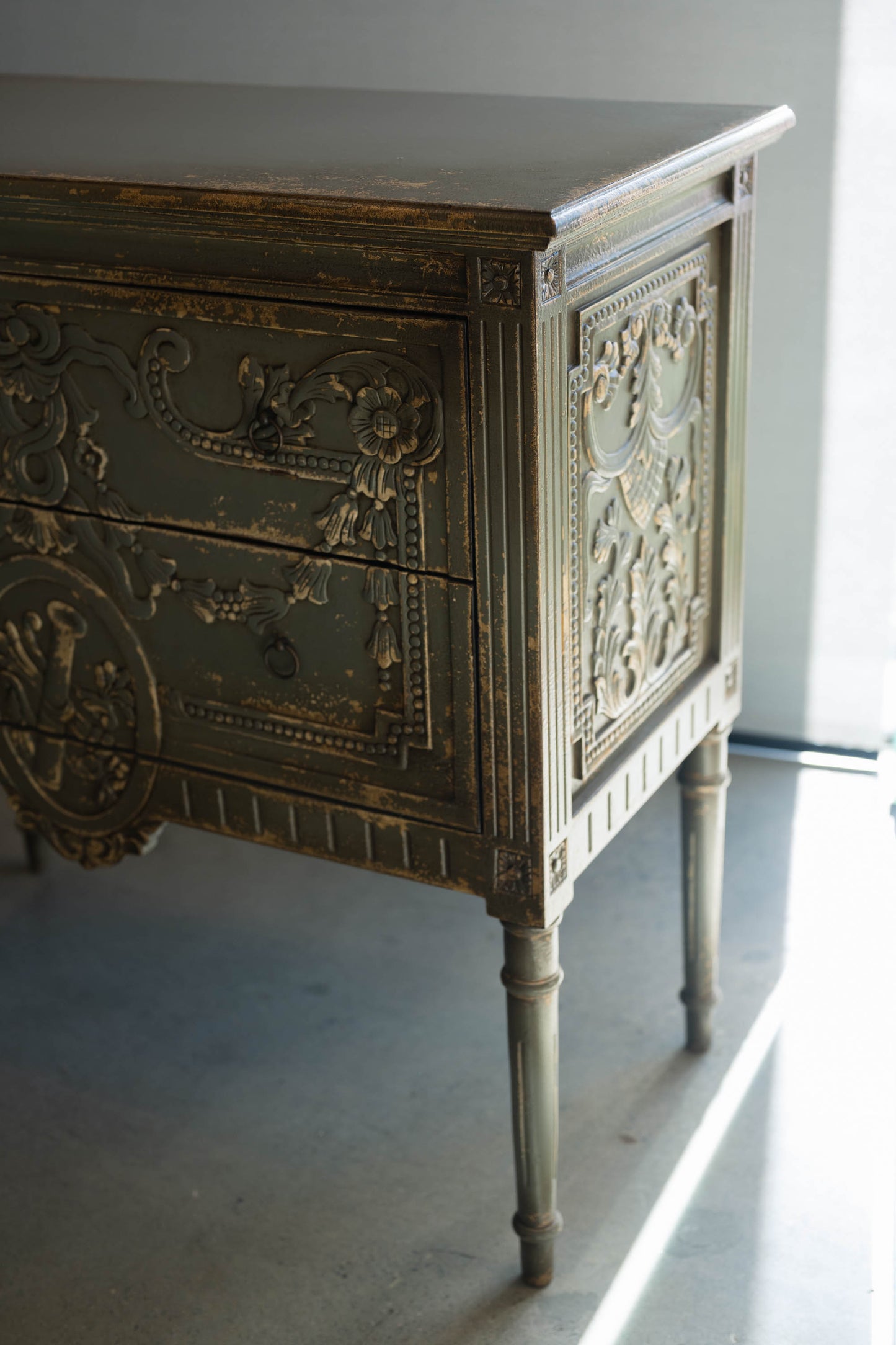 The unique and stunning one of a kind Daphne Sideboard boasts intricate carvings and distressed pale olive/grey painted finish. Crafted from mahogany and featuring two drawers, this versatile piece is perfect for a living or dining room in even in a bedroom. Corner