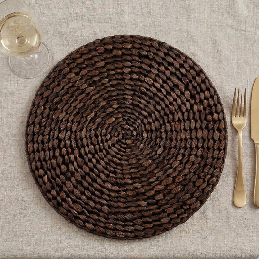 Round woven placemat on a table with gold cutlery and a glass of wine.