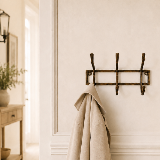 A rustic three-hook wall coat rack in a dark bronze finish, mounted on a cream-colored wall with decorative molding. A light beige wool coat is hanging from the center hook.