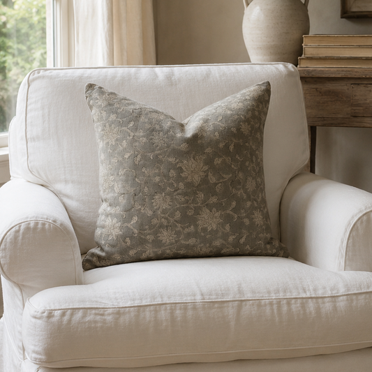 A white slipcovered armchair in a rustic living room setting, featuring a textured gray floral throw pillow. The chair is styled next to a wooden console table with a ceramic vase of greenery, a framed landscape print, and a woven basket.