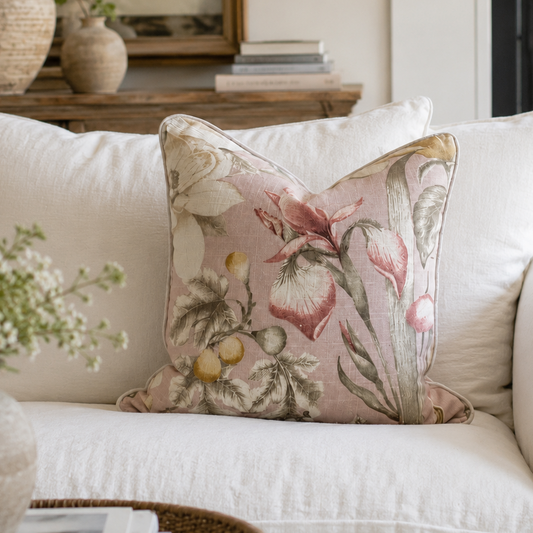 A square decorative throw pillow with a vintage floral and botanical pattern in muted pink, beige, and olive green, styled on a white linen sofa in a cozy living room setting.