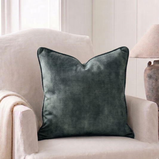 A square aqua green velvet throw pillow with piped edges, styled on a cream-colored armchair in a bright, modern living room setting.