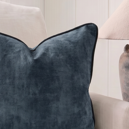A square navy blue velvet throw pillow with piped edges, styled on a cream-colored armchair next to a rustic table lamp.
