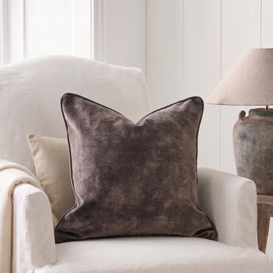 A square throw pillow in a textured, mottled dark brown or charcoal gray velvet fabric, styled on a cream-colored armchair next to a neutral lamp.