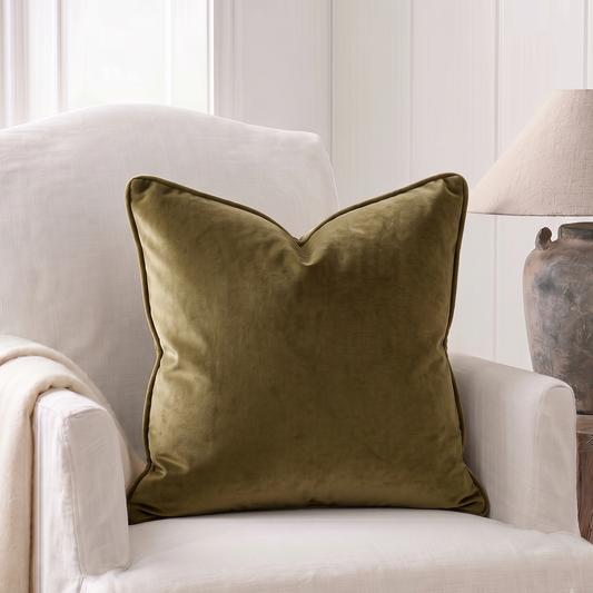 An olive green velvet throw pillow with a piped edge, styled on a cream-colored armchair next to a rustic table lamp.