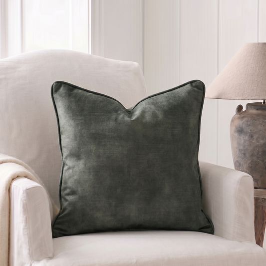 A square dark green velvet throw pillow with piped edges, styled on a cream-colored armchair next to a rustic table lamp.