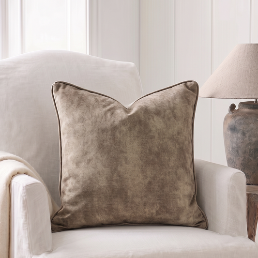 A square throw pillow in a textured taupe-brown velvet fabric with a subtle darker piped edge, styled on a cream-colored armchair next to a neutral throw blanket and a rustic table lamp.