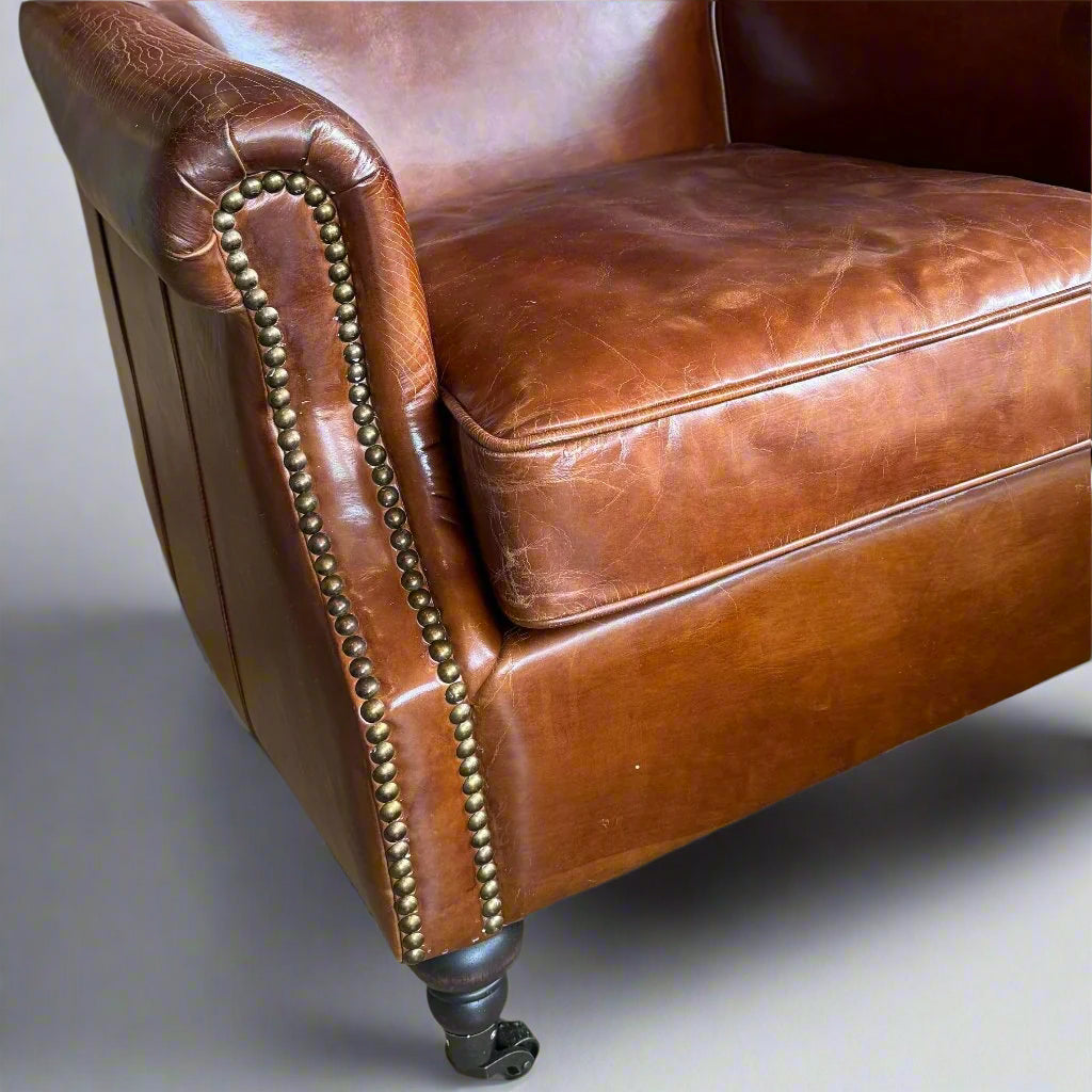 The Oliver Aged Leather Armchair boasts top grain cowhide upholstery, expertly aged by hand for a timeless and vintage appearance. Finished with charming brass stud accents and castor wheel feet. Detail