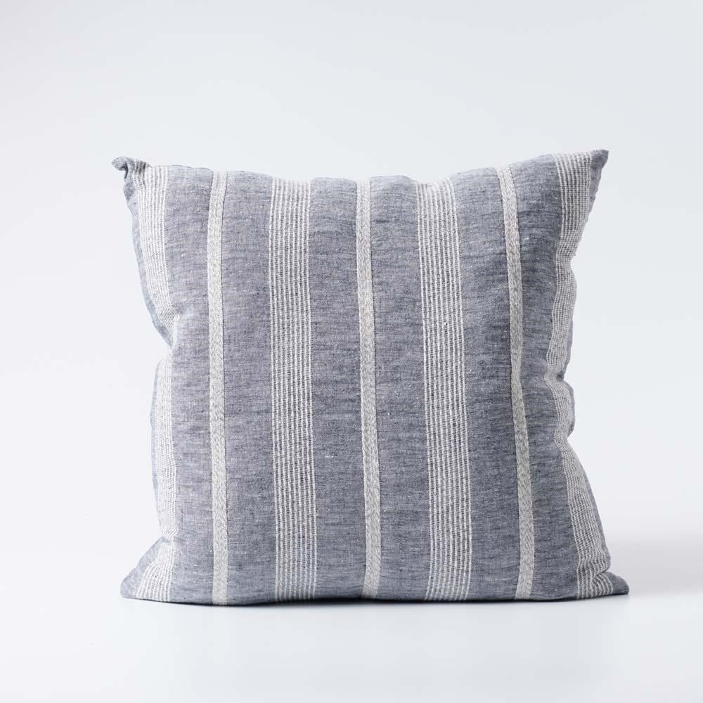 The Paros Chambray Striped Linen Cushion is made from a striking yarn-dyed chambray linen fabric featuring an elegant white stripe pattern. It comes with a superior plush feather insert, crafted to provide maximum comfort. Additionally, this product has attained OEKO-TEX® and GOTS certifications. Front.