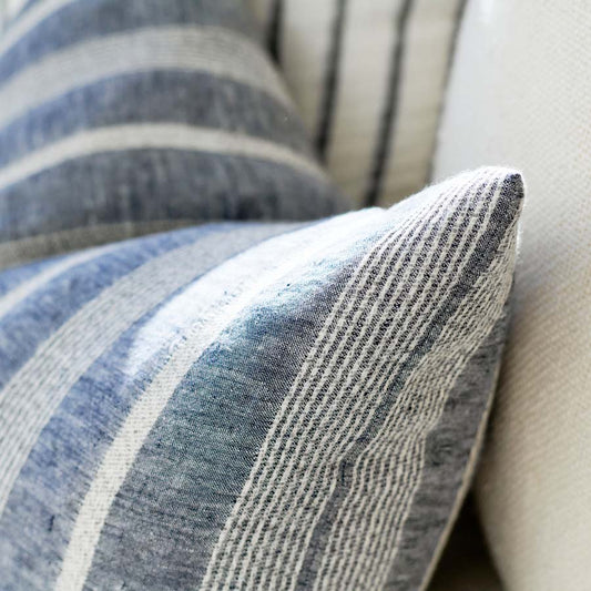 The Paros Chambray Striped Linen Cushion is made from a striking yarn-dyed chambray linen fabric featuring an elegant white stripe pattern. It comes with a superior plush feather insert, crafted to provide maximum comfort. Additionally, this product has attained OEKO-TEX® and GOTS certifications. Corner close up.