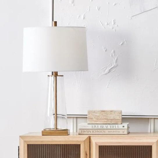 Experience the beauty of skilled craftsmanship with the Tivoli Glass Table Lamp. The clear glass body is accented by a hand-painted antique gold rod and plinth, adding a touch of elegance to any room. The off-white tapered linen shade adds to the delicate aesthetic, making it a truly stunning piece for your home.