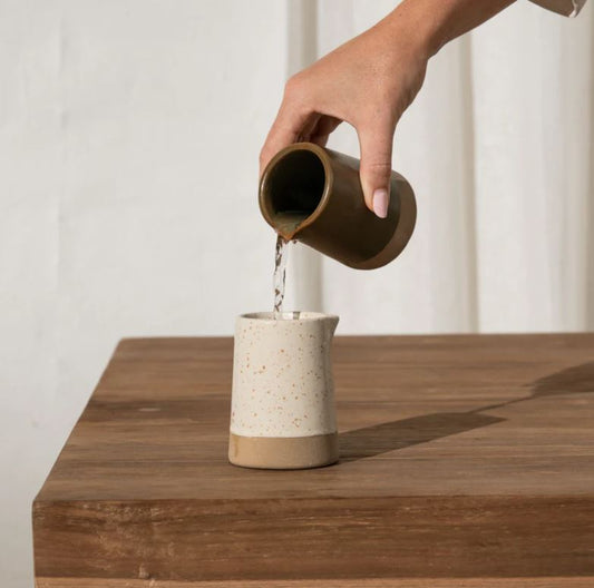 Person pouring water from a ceramic pitcher into another on a wooden surface.