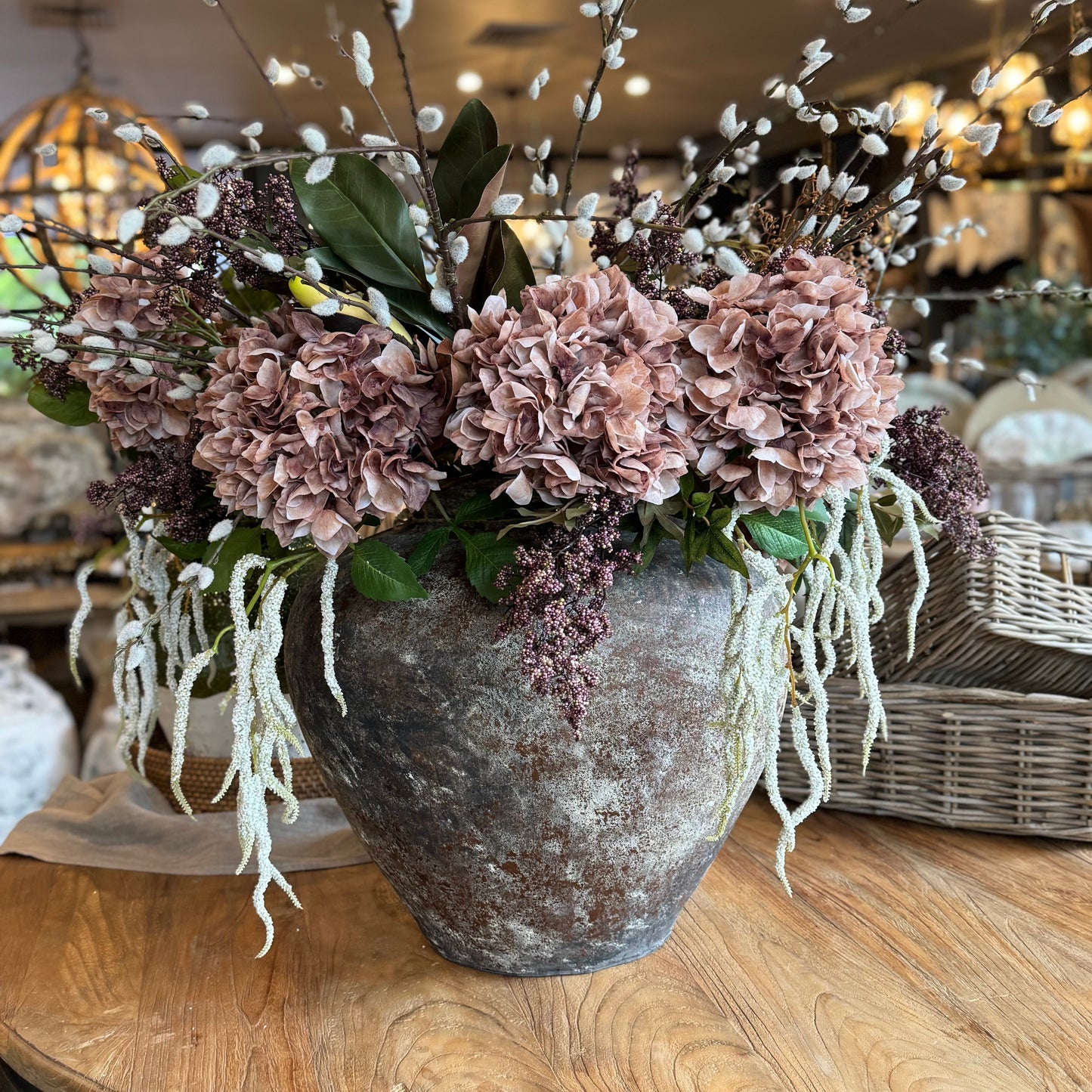 Decorative floral arrangement in a textured vase on a wooden table with a blurred indoor background.