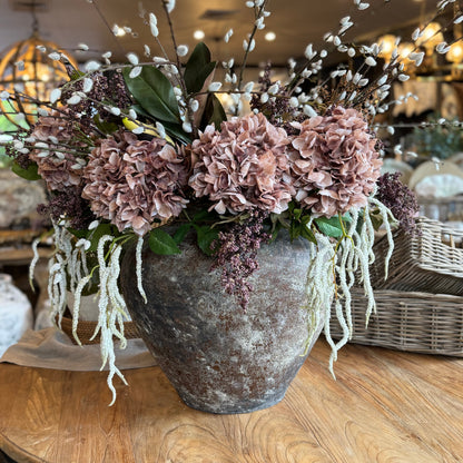 Decorative floral arrangement in a textured vase on a wooden table with a blurred indoor background.