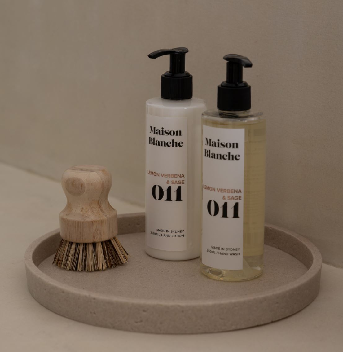 Two bottles of Maison Blanche hand lotion on a round stone tray with a wooden brush.