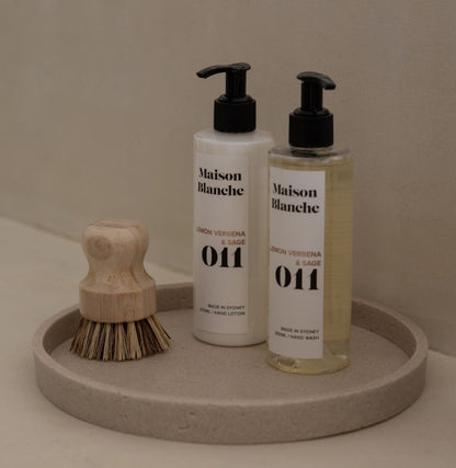 Two bottles of Maison Blanche hand lotion on a round stone tray with a wooden brush.