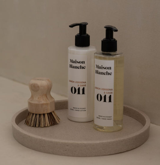 Two bottles of Maison Blanche hand lotion on a round stone tray with a wooden brush.