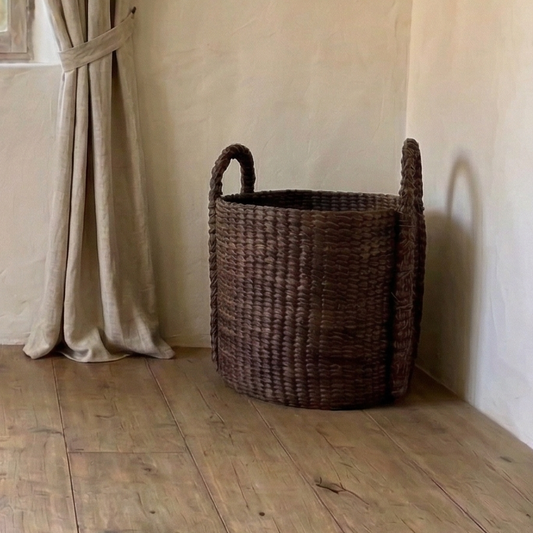Room with wooden floor, window with beige curtains, and a woven basket.