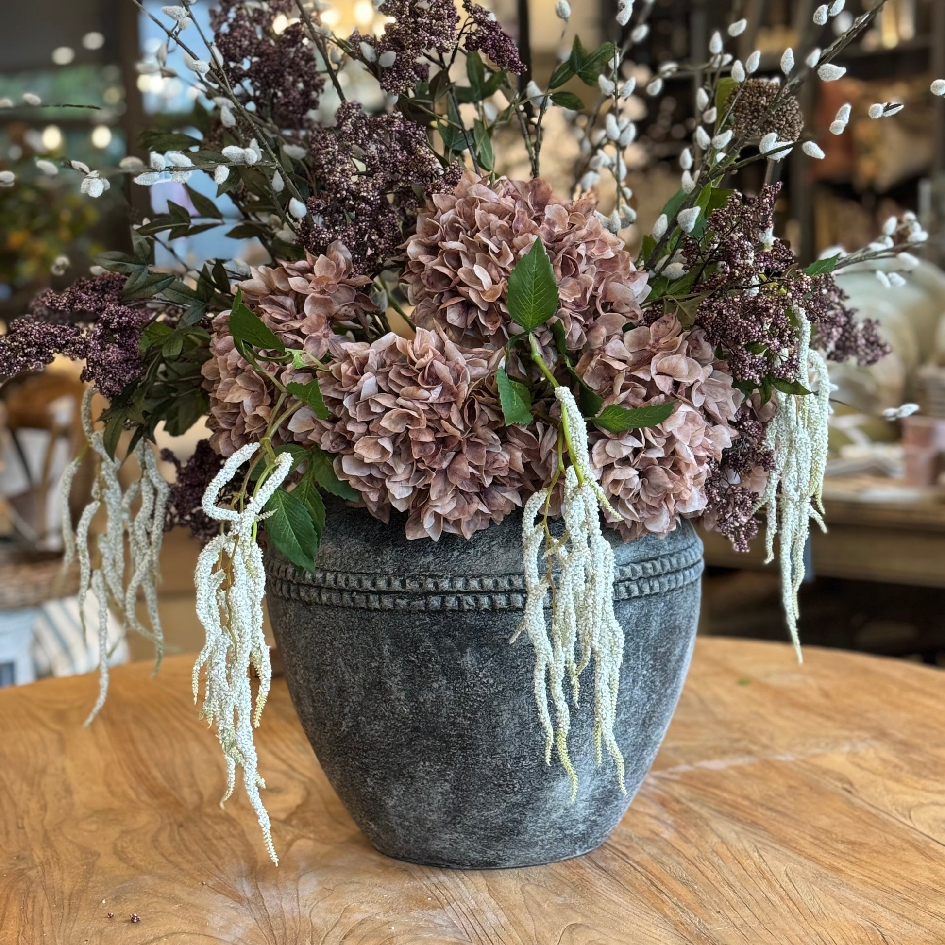 Decorative floral arrangement in a textured pot on a wooden table with a blurred indoor background.