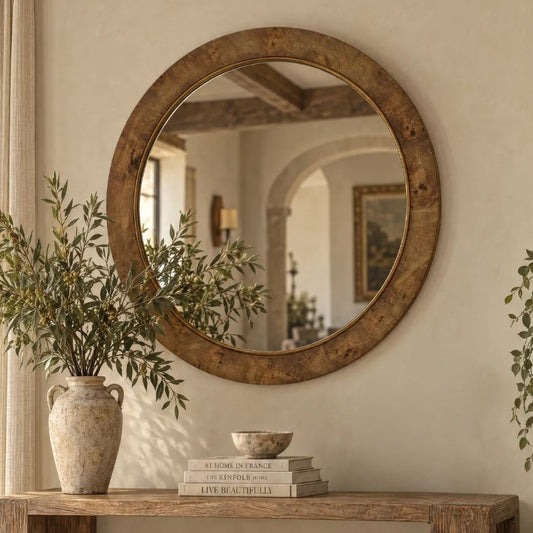 Round rustic wood-framed wall mirror hanging above a wooden console table with a vase of greenery and stacked books in a warm neutral interior.