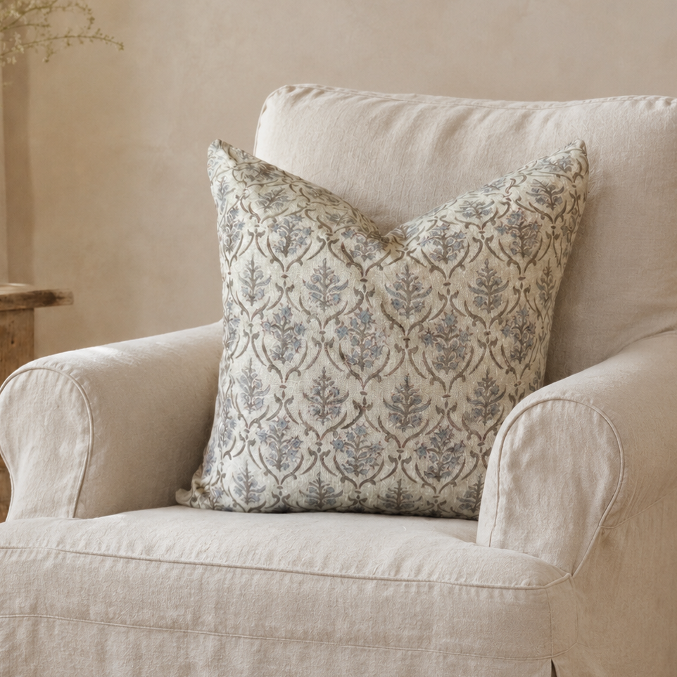 A cream-colored slipcovered armchair in a cozy room setting, styled with a square throw pillow featuring a blue-gray damask pattern on a beige background.