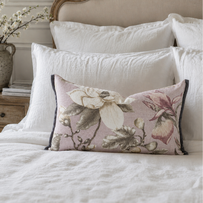 A rectangular floral throw pillow with a dusty pink background and cream, green, and mauve botanical print, styled on a bed with white linens and a beige upholstered headboard.
