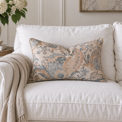 A rectangular decorative throw pillow with an intricate floral and botanical pattern in beige, blue, and gray, styled on a white linen sofa with a cream throw blanket.