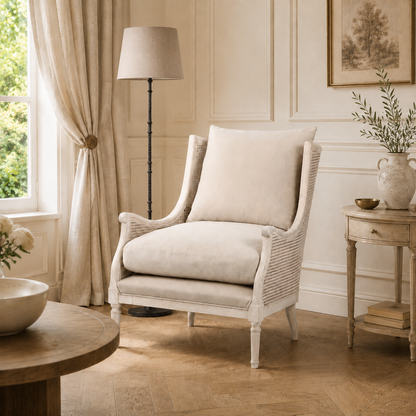 A classic white wicker armchair with plush cream cushions, styled in a bright, elegant room with beige walls, a floor lamp, and a side table.