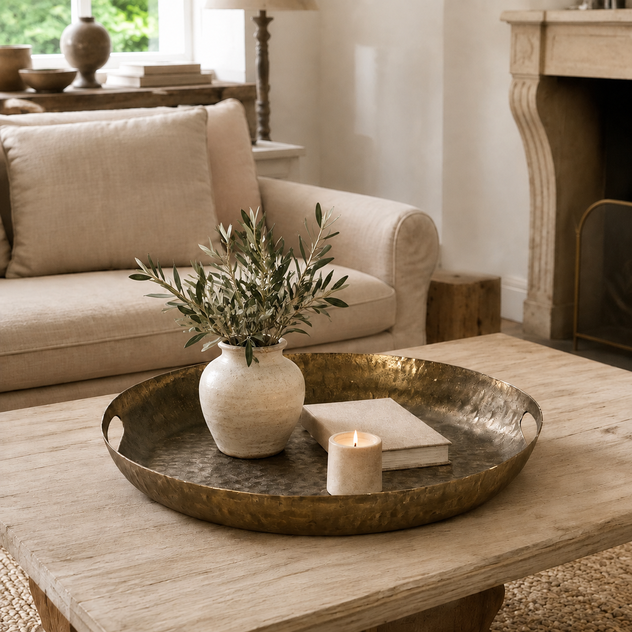 A hammered brass round tray styled on a rustic wooden coffee table in a cozy living room, holding a small beige ceramic vase with olive branches, a book, and a lit candle.