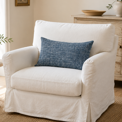 A white slipcovered armchair with a blue textured throw pillow, set in a bright, neutral-toned living room next to a wooden side table.