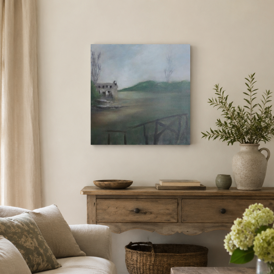 A square canvas painting of a serene lake landscape with a small stone cottage, displayed on a beige wall in a styled living room above a wooden console table.