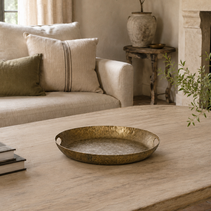 A round hammered brass tray with integrated side handles sits on a light wood coffee table in a beautifully styled living room with a beige sofa, fireplace, and potted plants.
