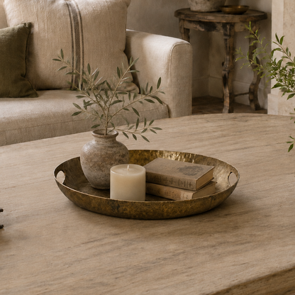 A styled living room scene featuring a light beige upholstered sofa with matching pillows, a rustic wooden coffee table, a brass tray with a candle and books, and potted plants near a stone fireplace.