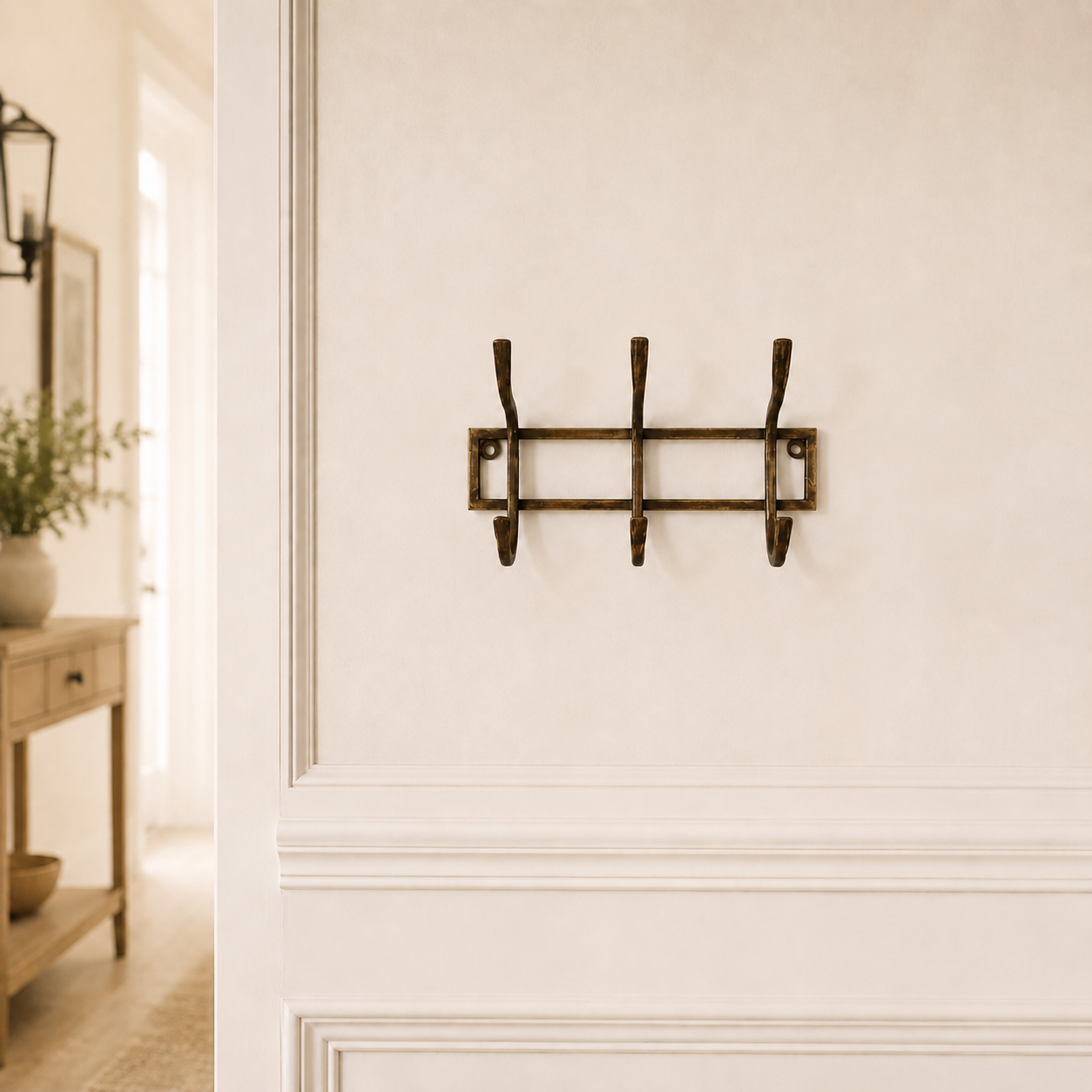 A three-hook wall hook rack in a dark bronze finish, mounted on a warm beige wall with decorative molding. The background shows a softly blurred interior hallway with a console table and lamp.