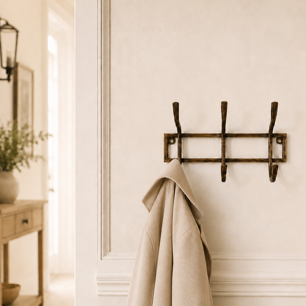 A rustic three-hook wall coat rack in a dark bronze finish, mounted on a cream-colored wall with decorative molding. A light beige wool coat is hanging from the center hook.