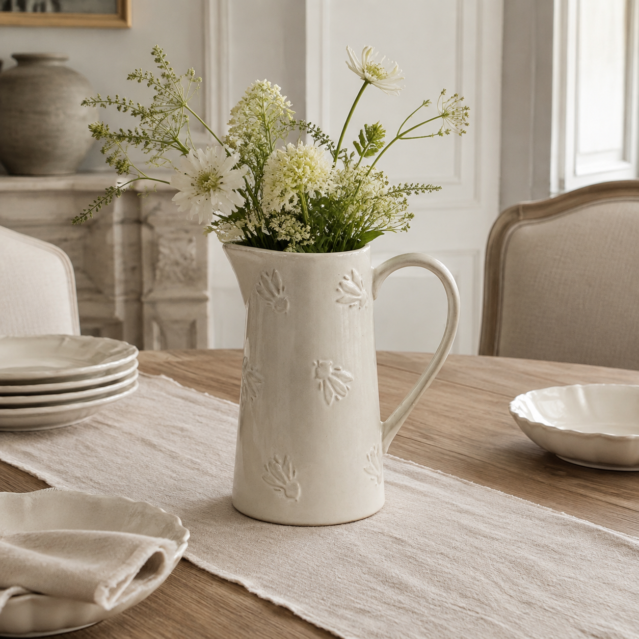 An off-white ceramic pitcher vase with embossed leaf patterns, filled with white and green wildflowers, set on a linen table runner on a wooden dining table.