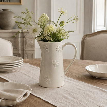 An off-white ceramic pitcher vase with embossed leaf patterns, filled with white and green wildflowers, set on a linen table runner on a wooden dining table.