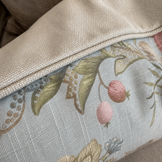 Close-up of a decorative throw pillow with a light blue floral and fruit print, featuring a beige woven fabric border, resting on a beige upholstered chair.
