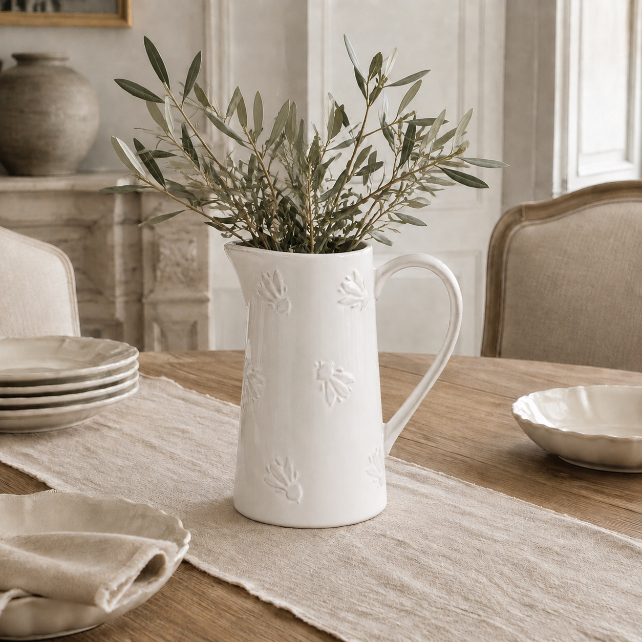 A white ceramic pitcher with embossed floral details, filled with olive branches, set on a wooden dining table with beige table linens and plates in a softly lit room.