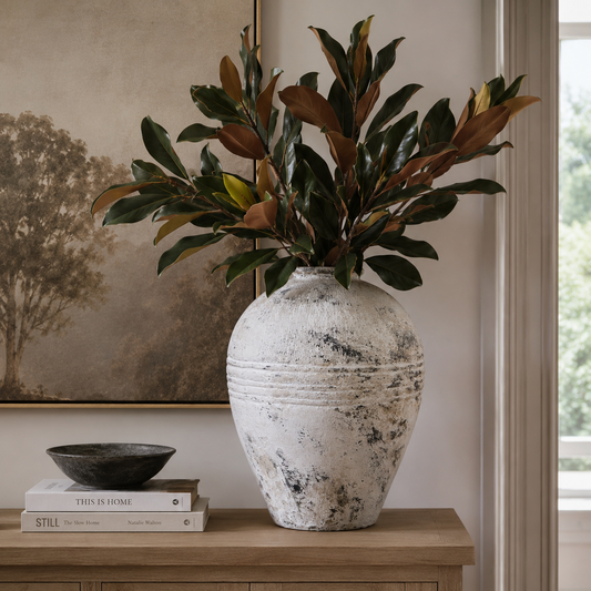 A large distressed white ceramic vase filled with green and brown magnolia leaves, styled on a light wood console table next to a window and a framed landscape painting.