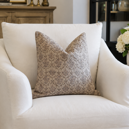 A white slipcovered armchair in a styled living room setting, featuring a textured beige and gray patterned accent pillow.