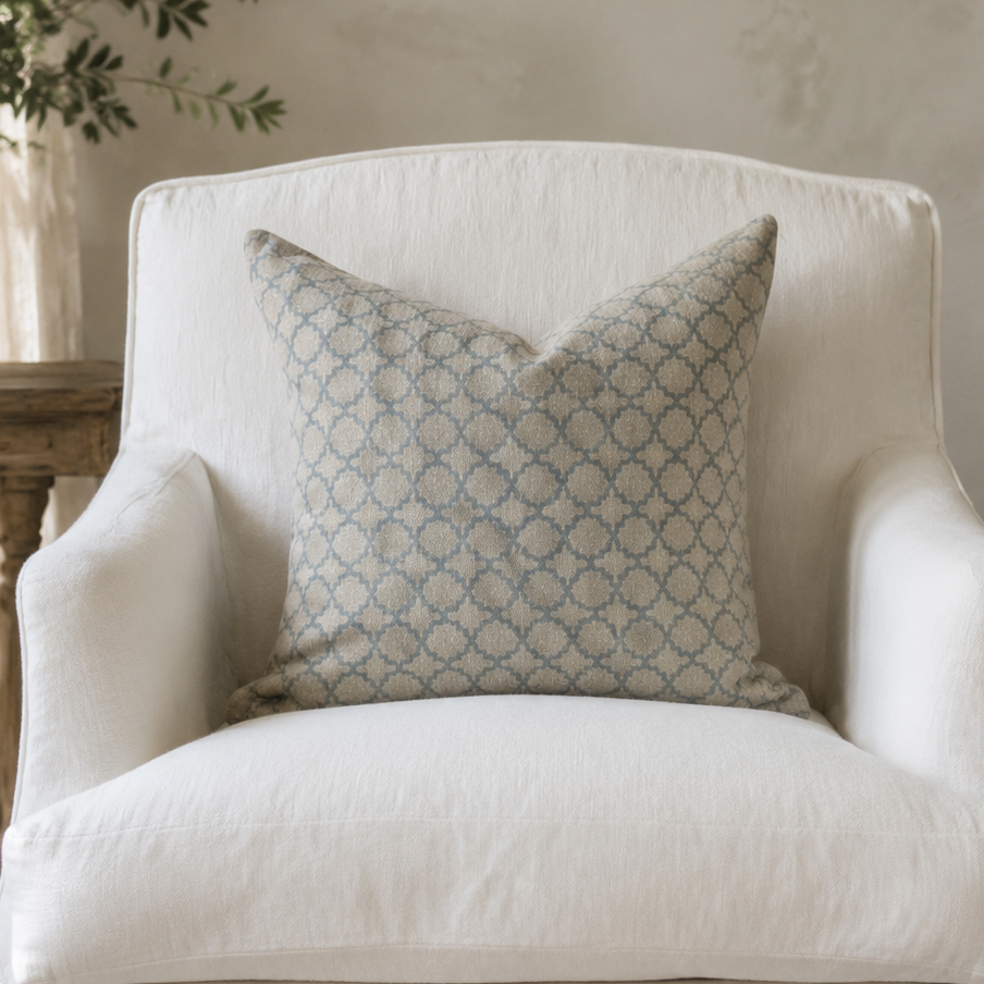 A classic French-style armchair upholstered in off-white linen fabric with carved light wood legs, styled with a blue and beige patterned accent pillow in a sunlit room.