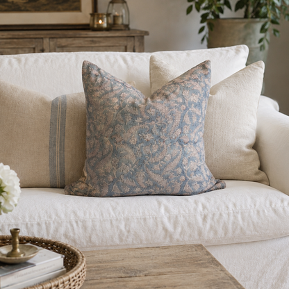A white slipcovered sofa in a bright living room, styled with multiple beige and blue patterned throw pillows. A wooden coffee table with a woven tray and white flowers sits in the foreground, and a window with sheer curtains is visible on the right.