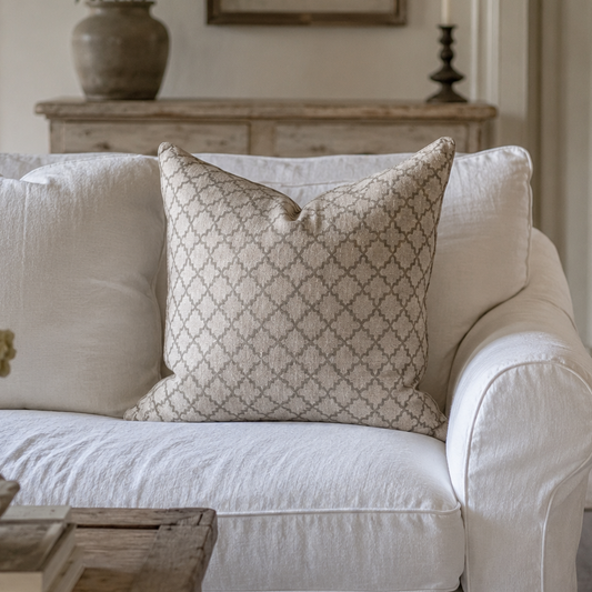 A beige throw pillow with a geometric trellis pattern rests on a white slipcovered sofa in a softly lit, rustic living room.