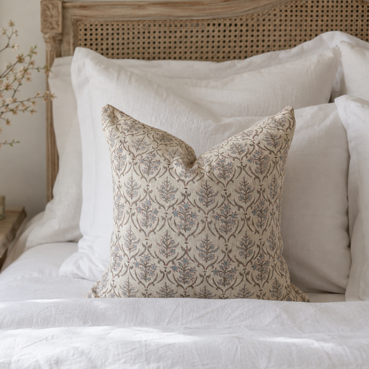 A decorative square throw pillow with a beige and muted blue-gray botanical damask pattern, styled on a white bed with a wooden cane headboard.