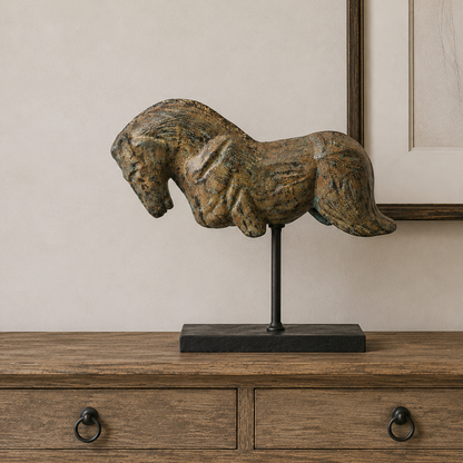 A rustic horse sculpture with a textured, weathered finish in earthy brown and gold tones, mounted on a black metal stand. The sculpture is displayed on a wooden console table against a neutral beige wall with framed artwork in the background.