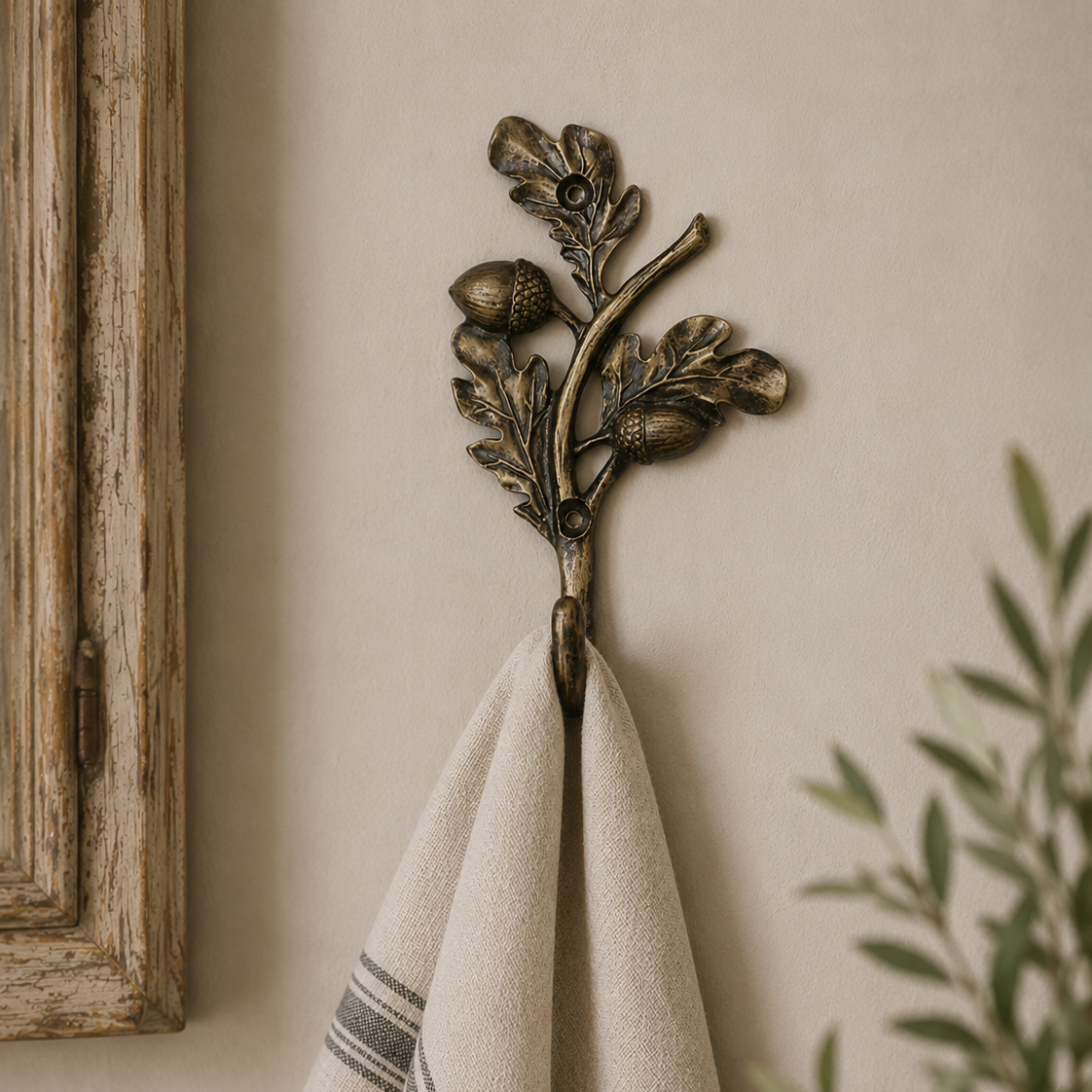 An antique bronze branch-shaped towel hook with acorns and leaves, hanging on a warm beige wall. A cream-colored linen towel with gray stripes is draped over the hook, with a rustic wooden frame on the left and a green potted plant on the right.