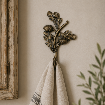 An antique bronze branch-shaped towel hook with acorns and leaves, hanging on a warm beige wall. A cream-colored linen towel with gray stripes is draped over the hook, with a rustic wooden frame on the left and a green potted plant on the right.