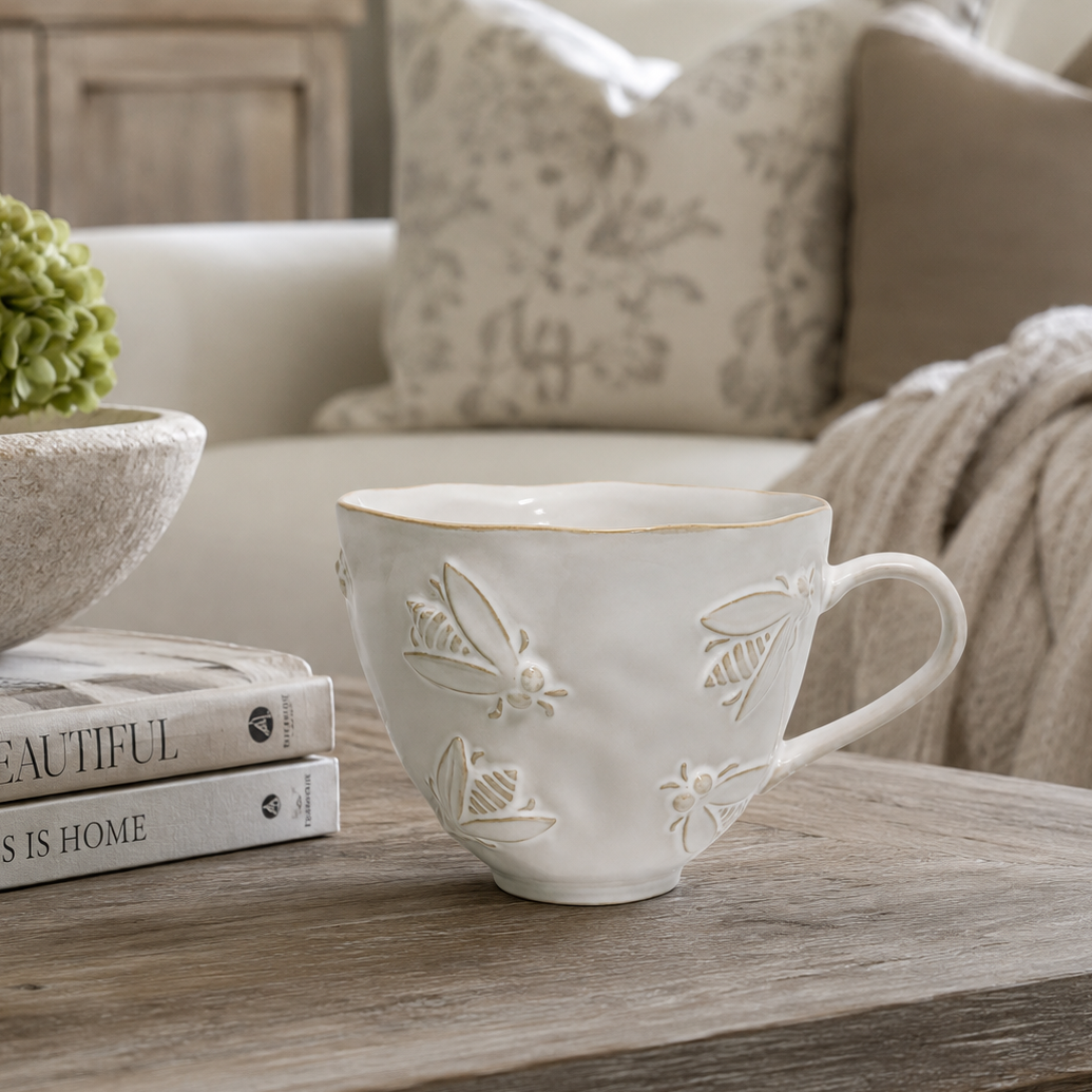 A white ceramic teacup with embossed floral and butterfly motifs sits on a rustic wooden table in a cozy living room setting, styled with books and a green hydrangea.